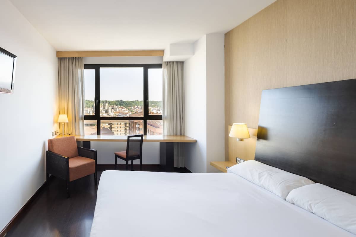 Une chambre d’hôtel avec un lit, un bureau et une chaise, une fenêtre avec des rideaux et une vue sur le paysage urbain.