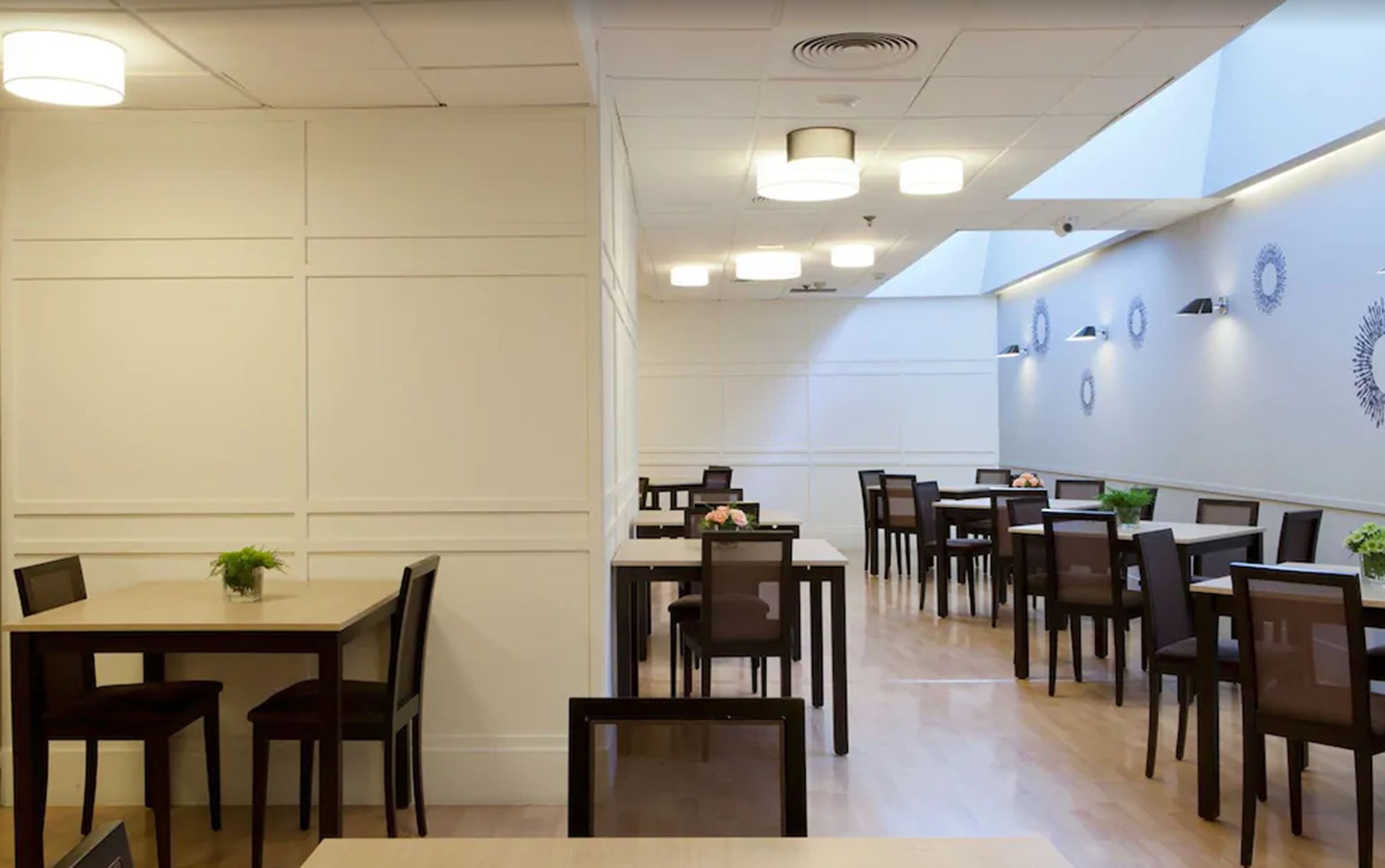 A dining area with multiple tables and chairs, white walls, and ceiling lights.