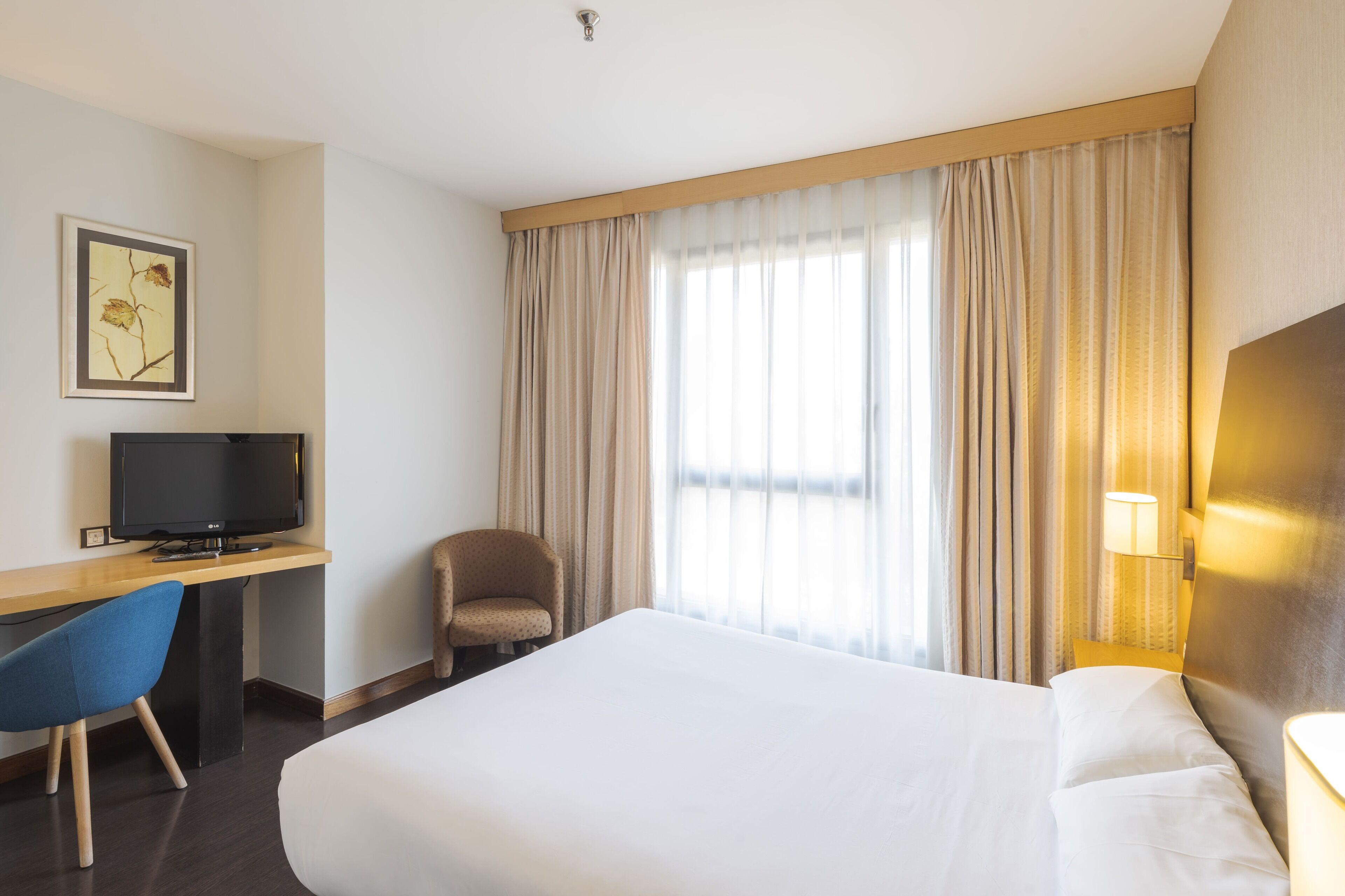A hotel room with a large bed, a desk with a television, a chair, and a window with curtains.