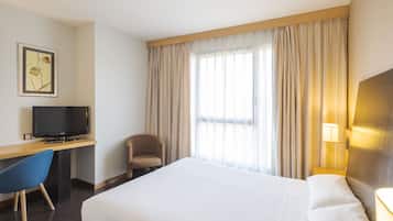 A hotel room with a large bed, a desk with a television, a chair, and a window with curtains.