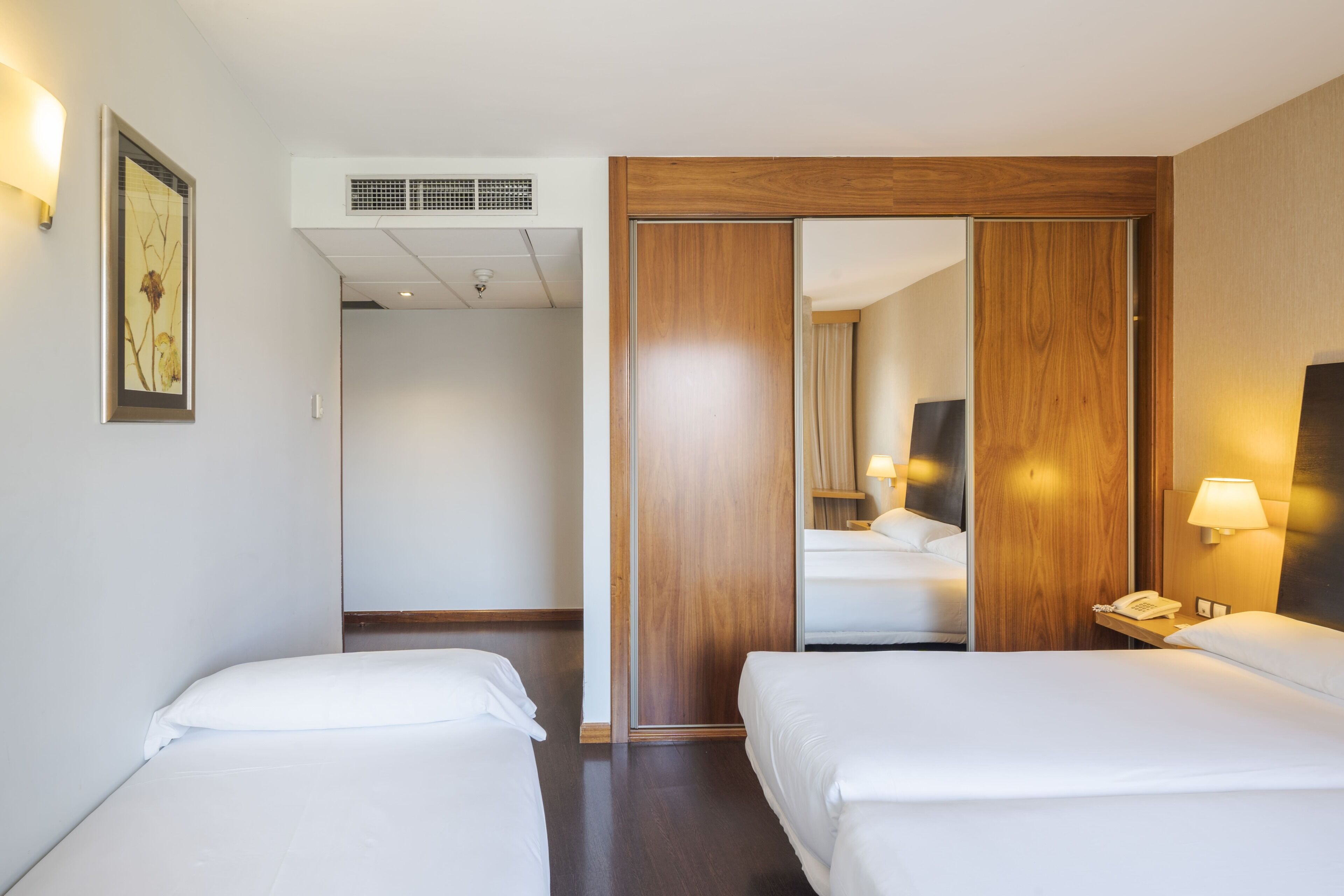 Habitación de hotel con dos camas, un armario de madera, un espejo, una lámpara de noche y un cuadro en la pared.