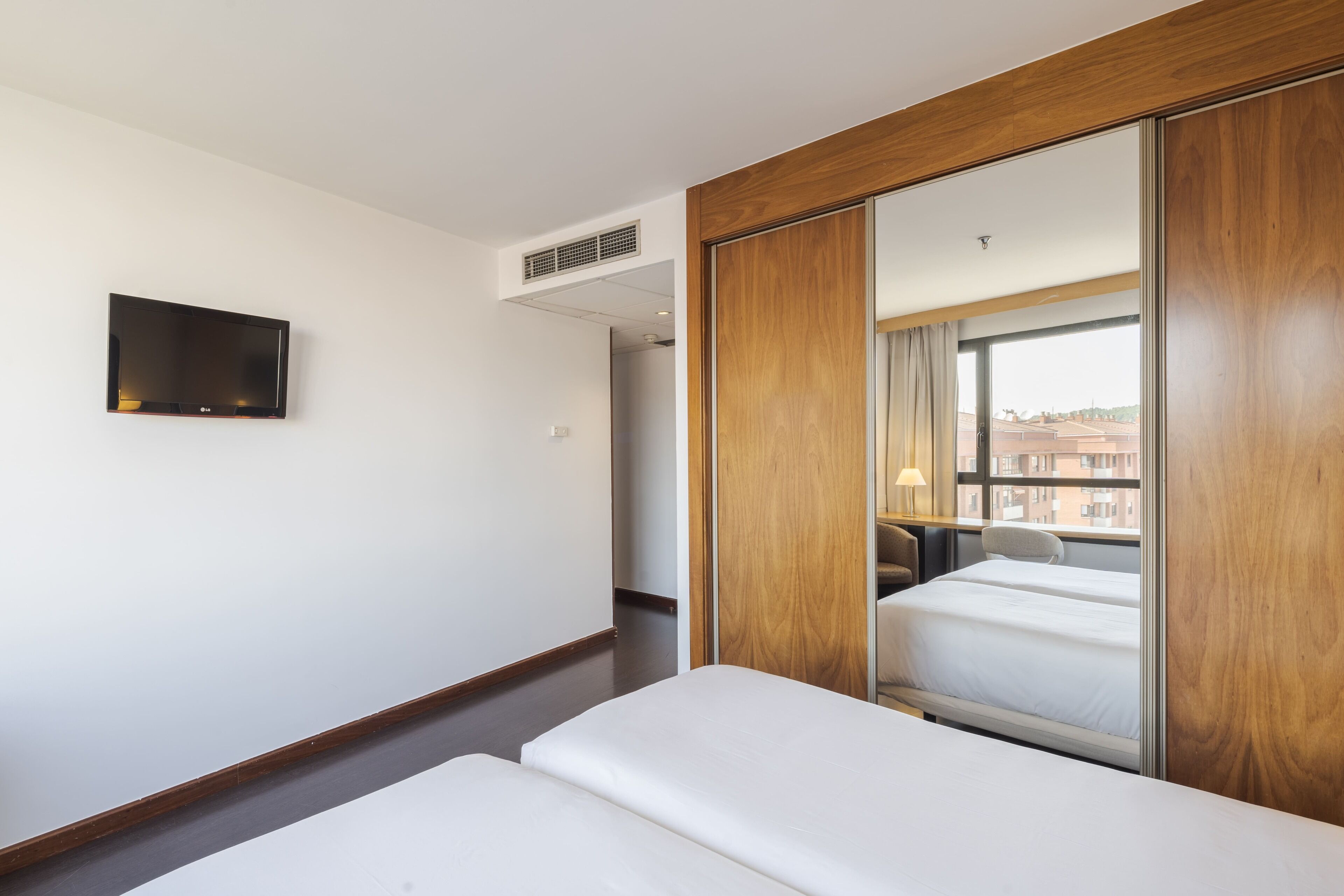 A hotel room with a bed, a TV mounted on the wall, and a large mirror reflecting the room.