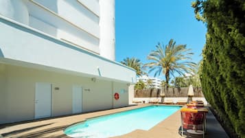 A swimming pool area with lounge chairs and a lifebuoy, surrounded by a white building and palm trees.