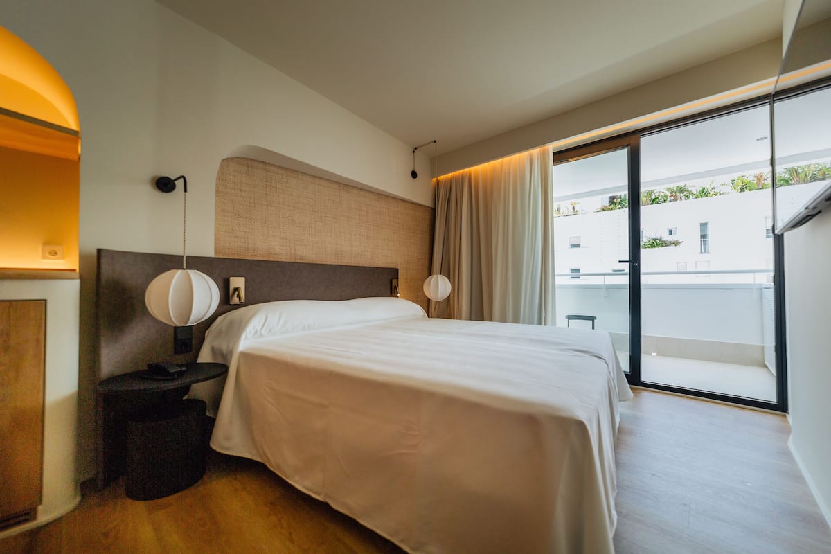 Una habitación de hotel moderna con una cama grande, una mesita de noche con lámpara, vistas al balcón exterior y una puerta corredera de cristal.