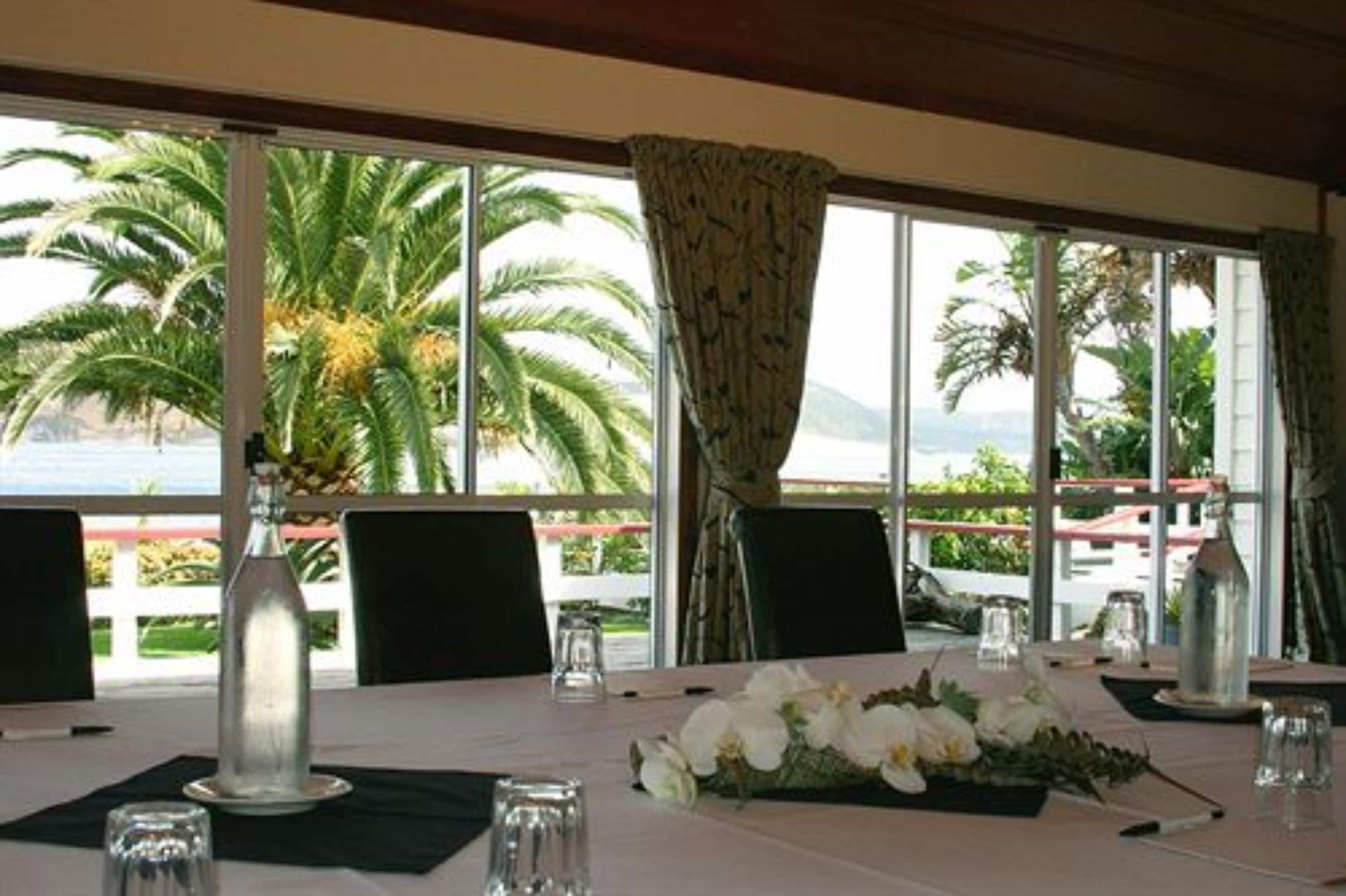 Un comedor con una mesa dispuesta para dos, ventanas amplias con cortinas, y vistas a palmeras y un edificio.