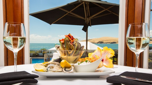 A table with seafood dishes, two wine glasses, and a view of the ocean. - The Sands Hotel (Omapere)