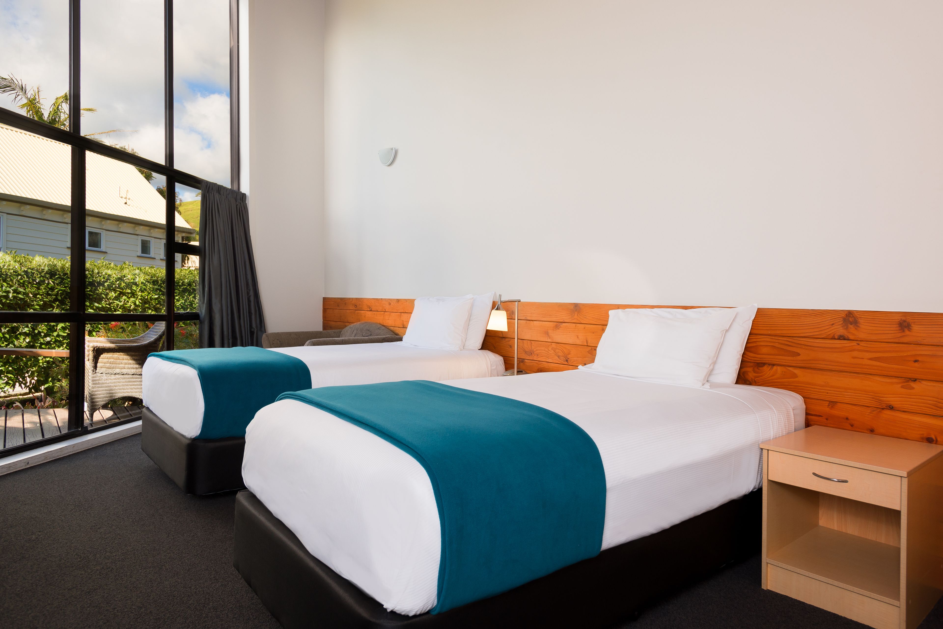 A hotel room with two beds, a wooden headboard, a small bedside table, and a view of greenery outside.