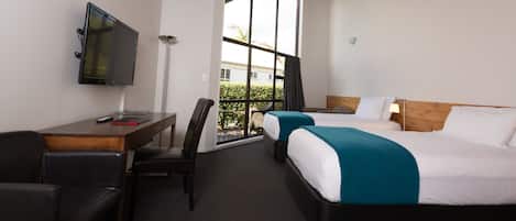 A hotel room with a wooden ceiling, a desk with a TV, two beds, and a view through a large window.