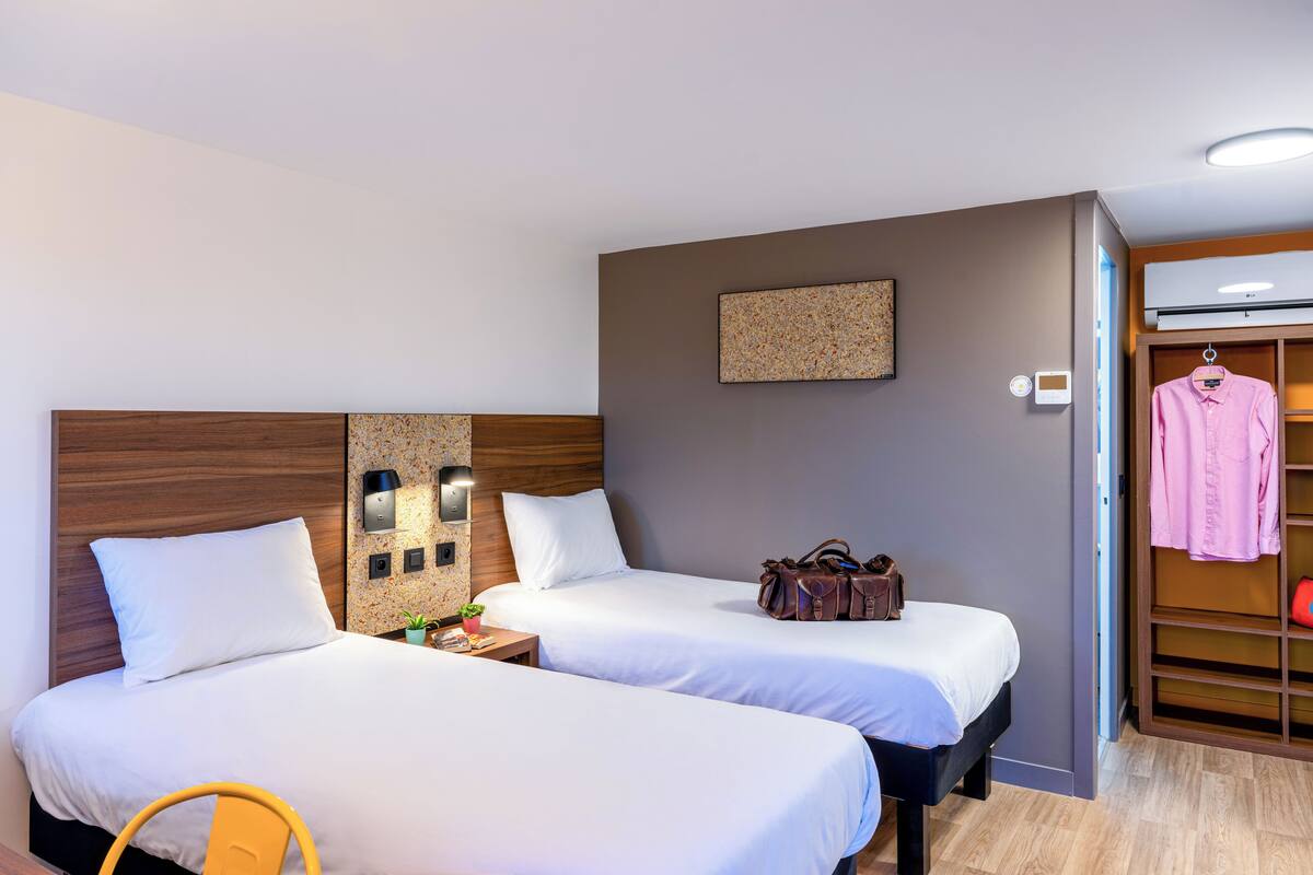 A hotel room with two beds, a wooden headboard, a small table with a plant, a wall-mounted artwork, and a wardrobe with hanging clothes.