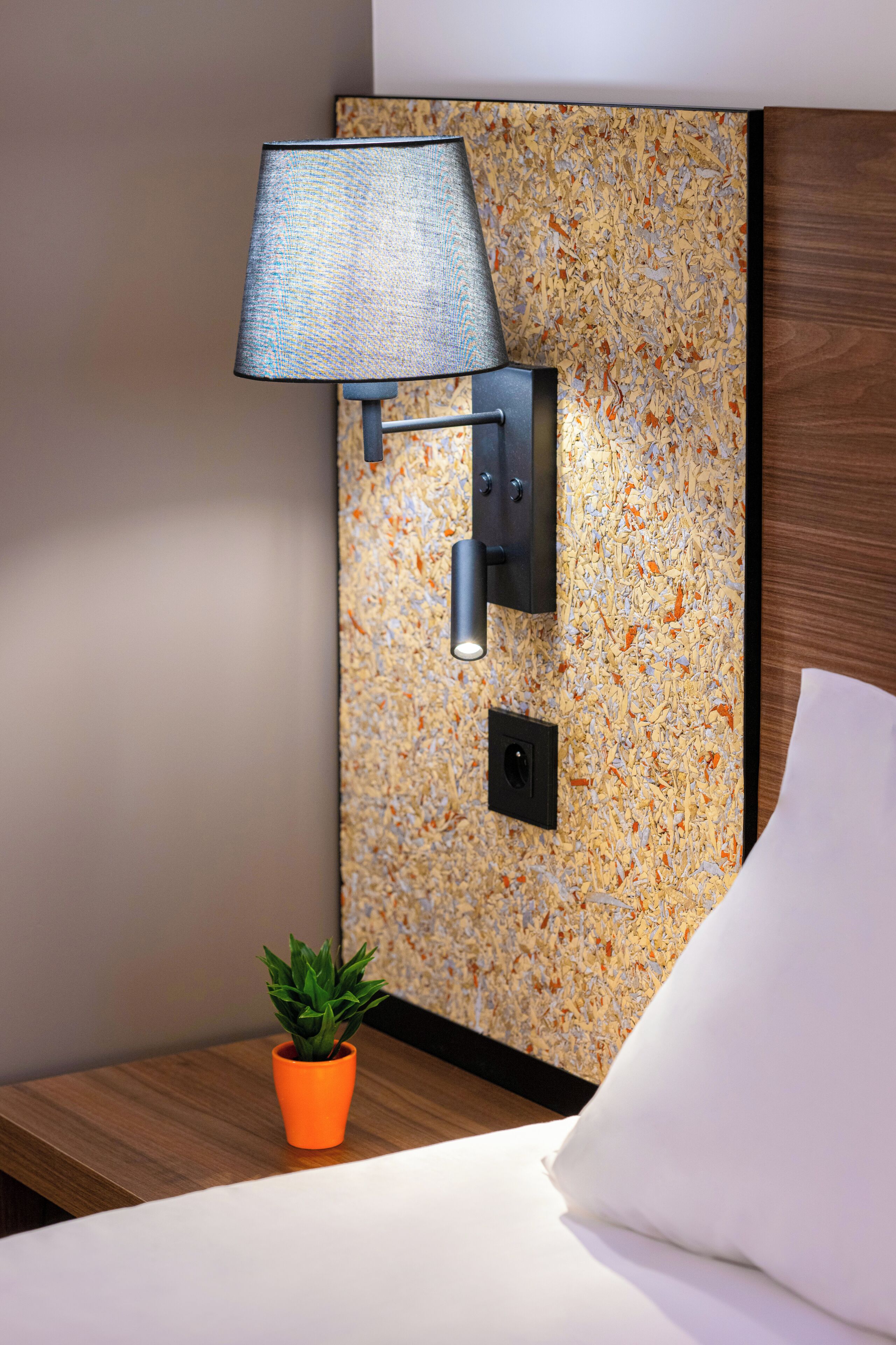 A modern hotel room with a textured wall, a wall-mounted lamp, a small potted plant, and a neatly made bed.