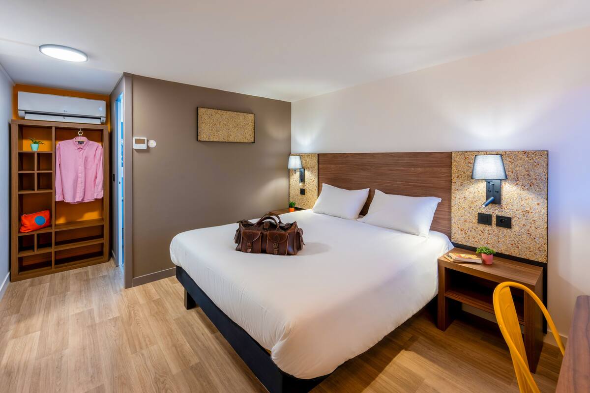 A neatly arranged hotel room with a large bed, bedside lamps, a wooden nightstand, a chair, a wardrobe, and a small table with a plant.