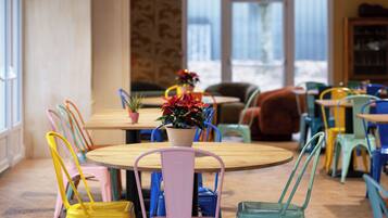 A dining area with colorful chairs and tables, a potted plant, and a view of the outdoors through large windows.