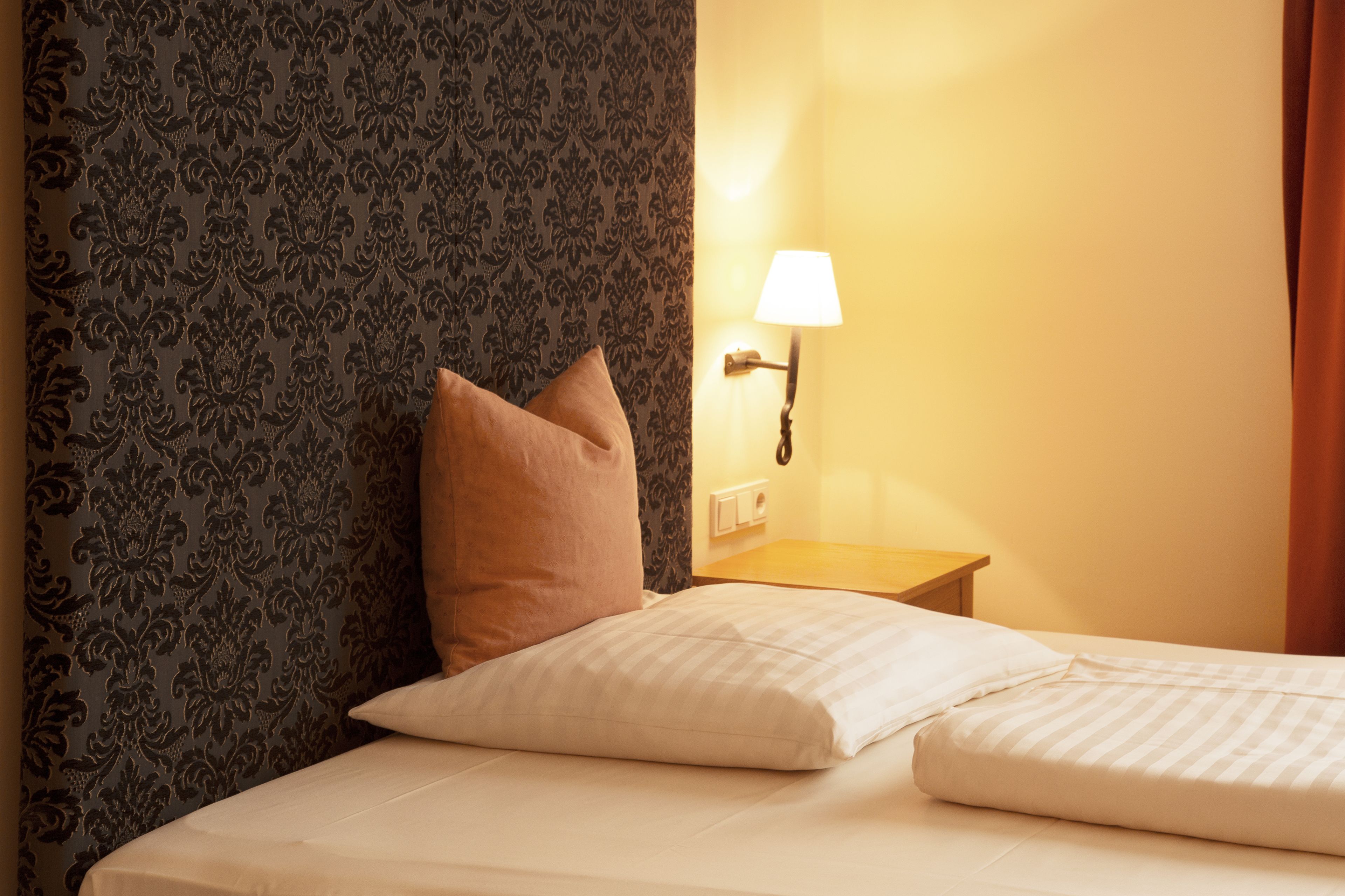 A neatly made bed with white linens and a single brown pillow, a wall-mounted lamp, and a patterned wallpaper.