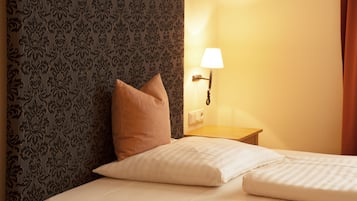 A neatly made bed with white linens and a single brown pillow, a wall-mounted lamp, and a patterned wallpaper.