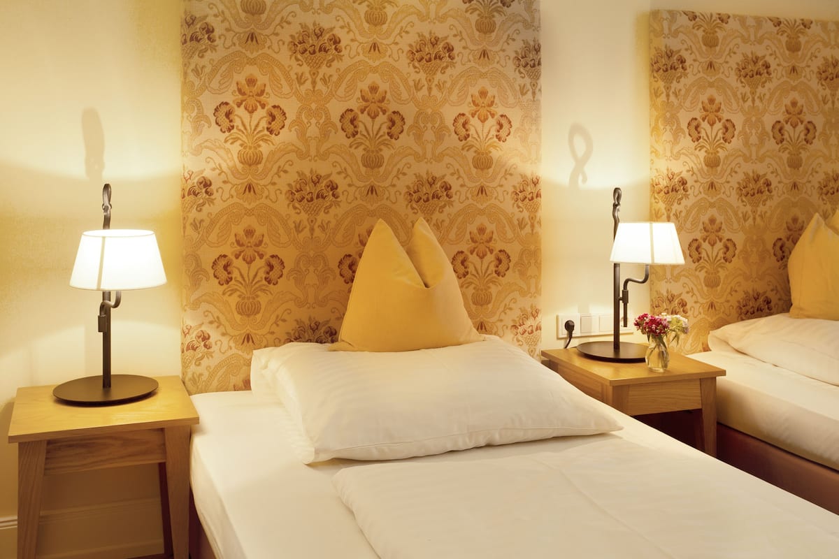 Quarto de hotel com duas camas, cada uma com mesa de cabeceira, luminárias e almofadas decorativas.
