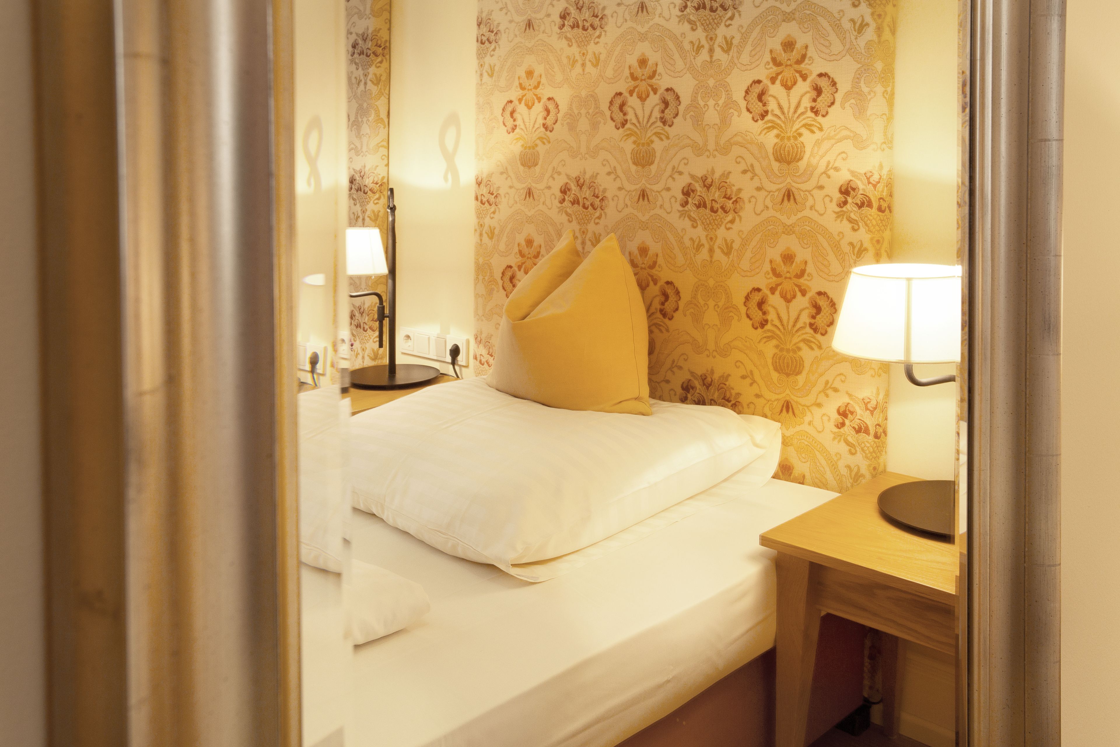 A hotel room with a bed, a bedside table with a lamp, and a patterned wallpaper.