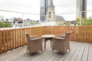 Una terraza de madera con muebles de mimbre y vista a un paisaje urbano que incluye una iglesia y edificios modernos.