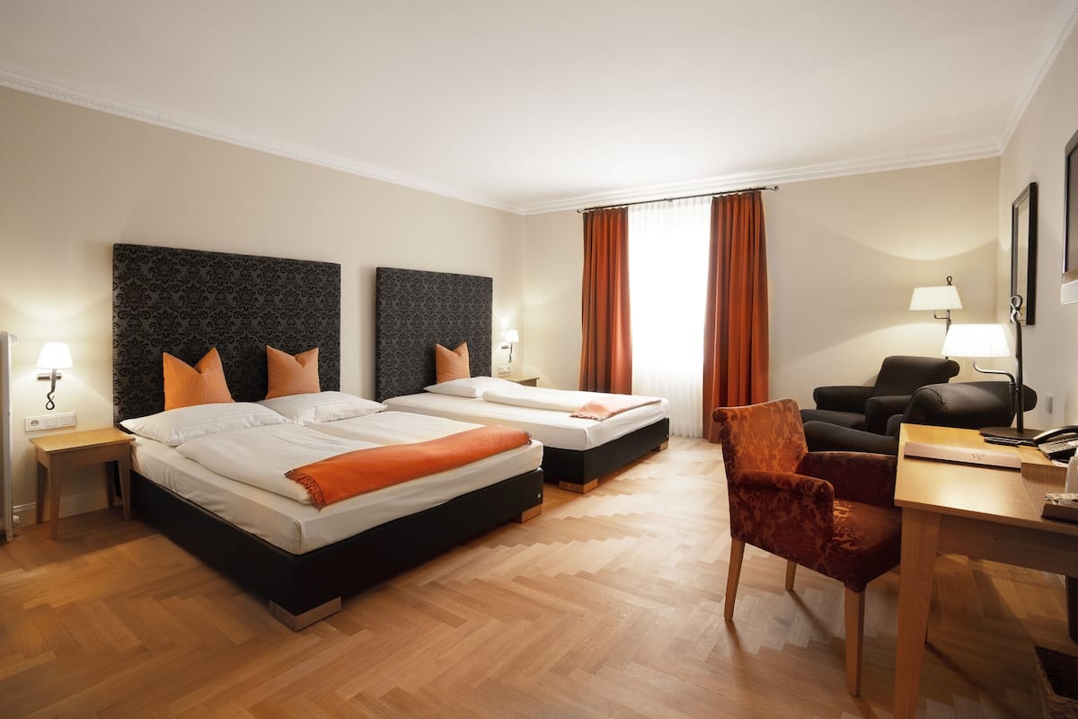 Quarto de hotel com duas camas, uma escrivaninha, uma cadeira e uma janela grande com cortinas.