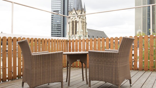 Une terrasse en bois avec des meubles en osier, une vue sur des immeubles modernes et, au loin, le clocher d’une église.