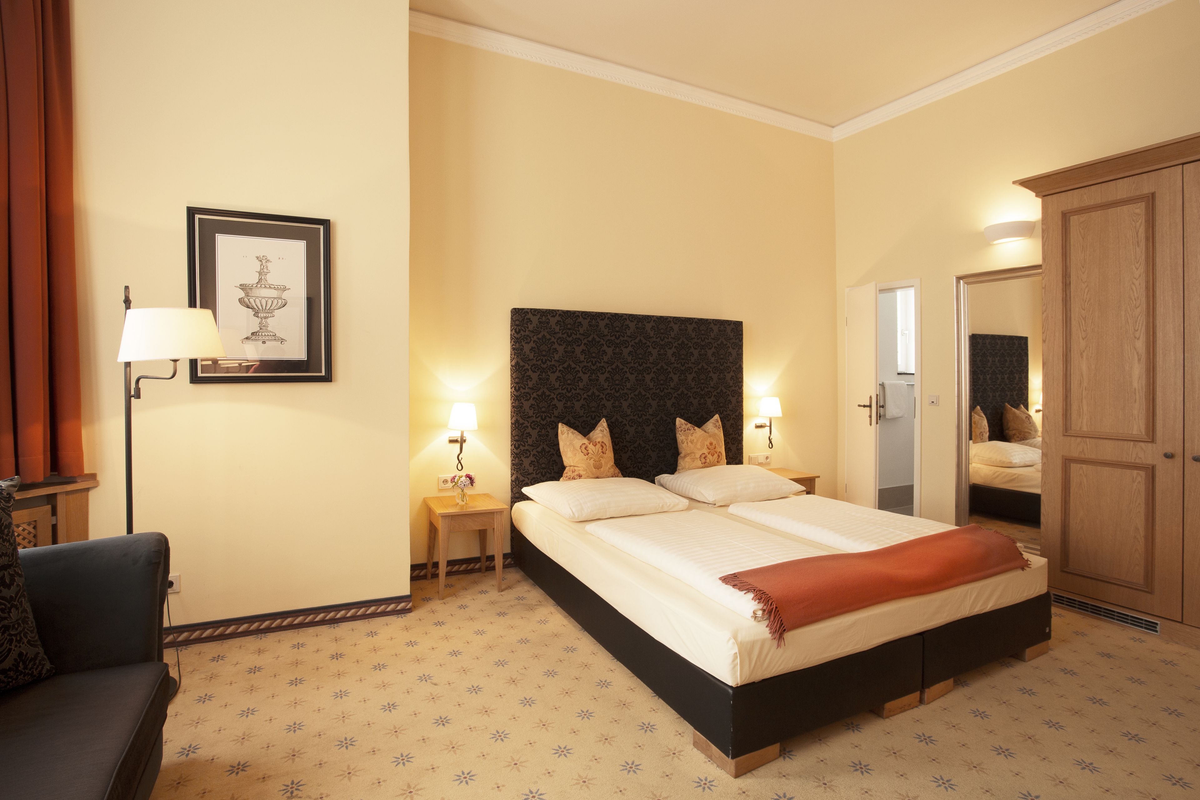 Une chambre d’hôtel avec un grand lit, des lampes de chevet, un tableau encadré au mur, une armoire en bois et une petite table avec une lampe.