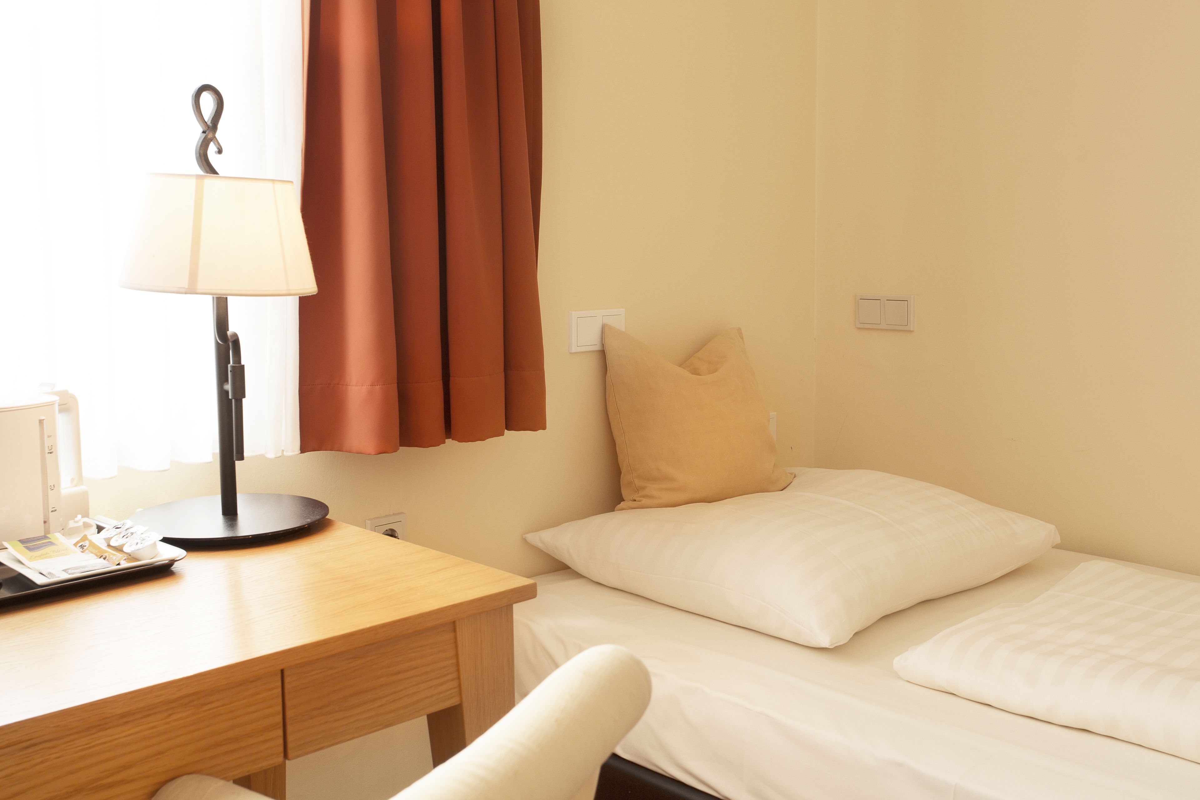 Habitación de hotel con un escritorio de madera, una lámpara, una cama con dos almohadas y una ventana con cortinas.