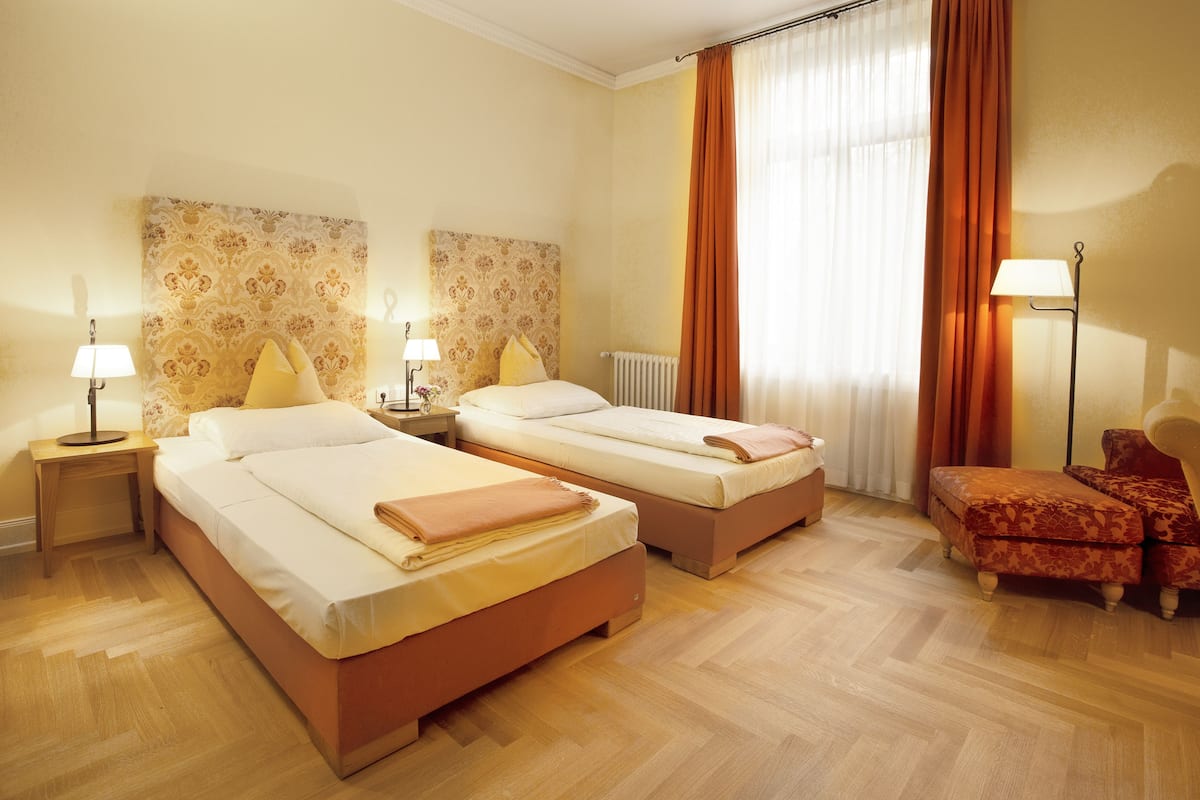 Quarto de hotel com duas camas, uma mesa de cabeceira com abajur, uma cadeira e uma janela grande com cortinas.