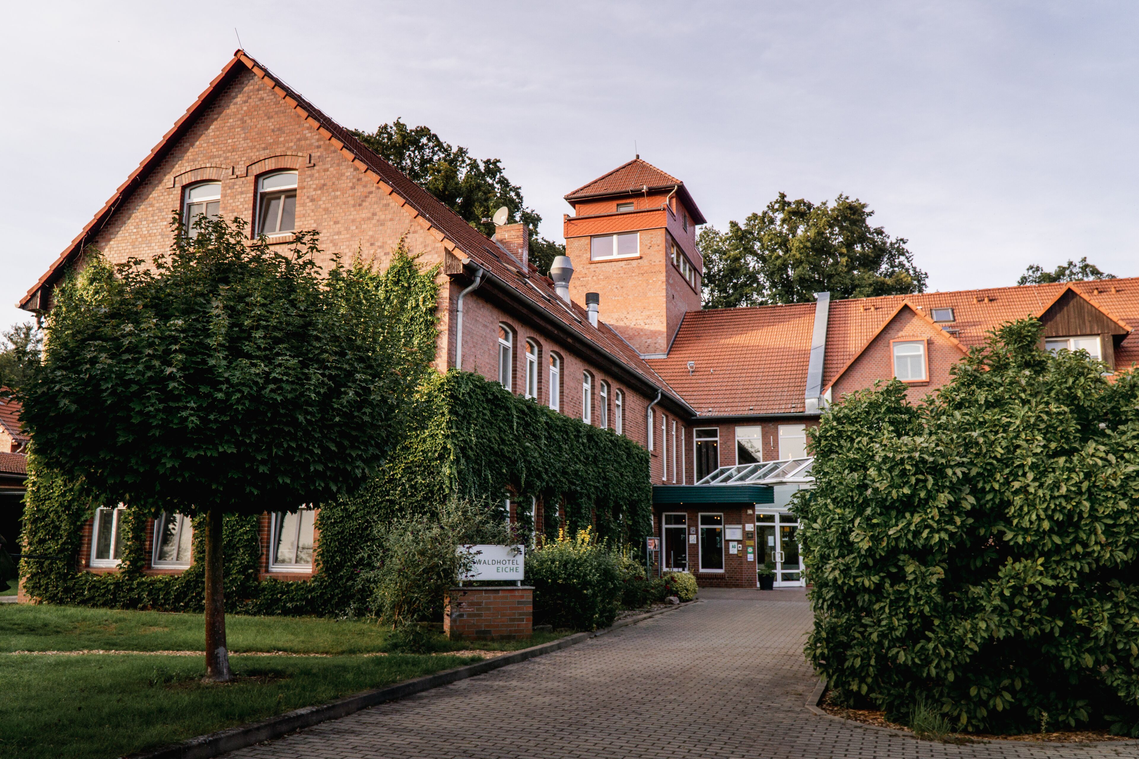 Ein traditionelles Backsteingebäude mit einem zentralen Turm, einer gepflasterten Auffahrt und umliegender Bepflanzung.