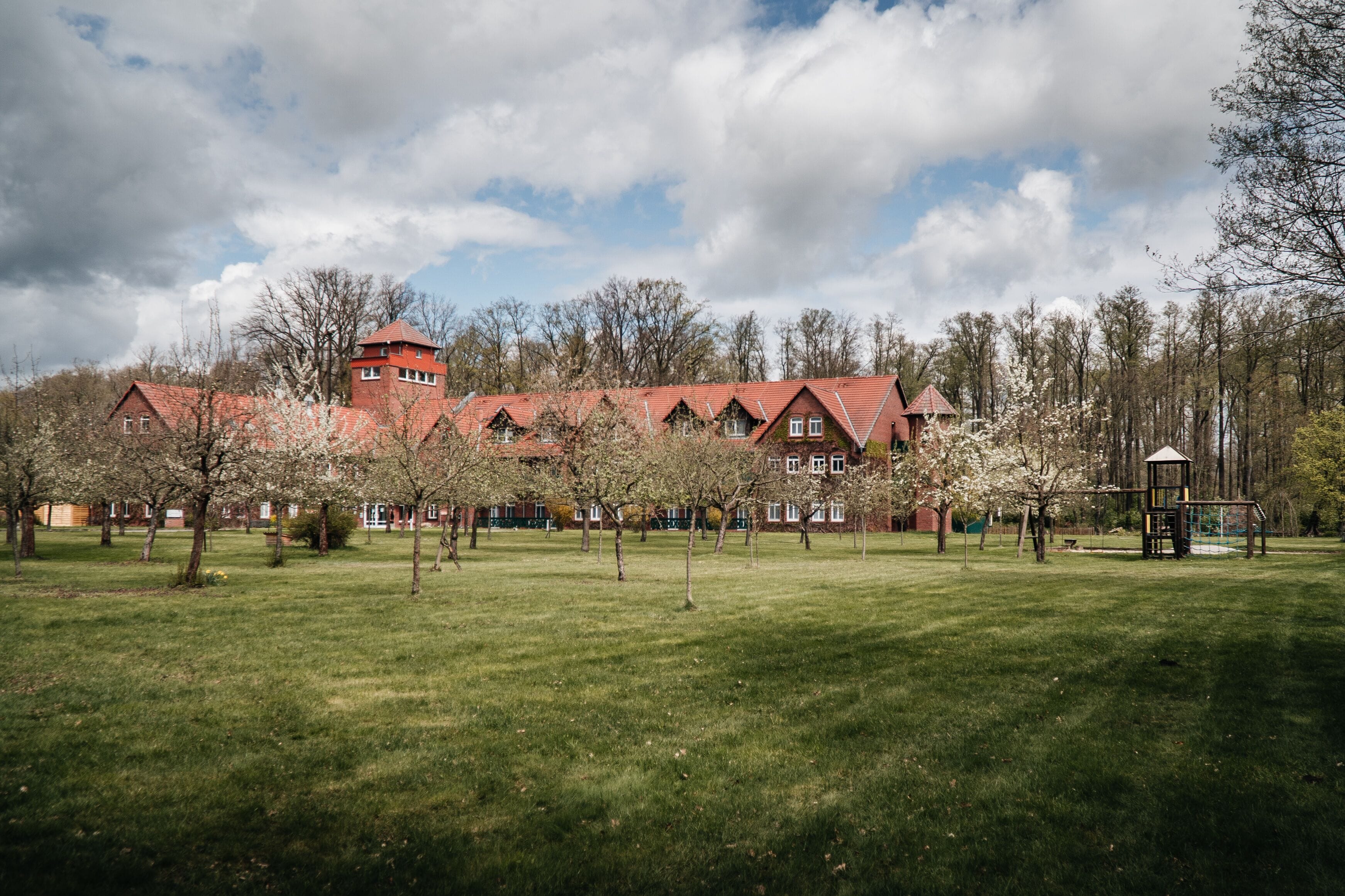 A large building with a red roof and multiple windows, surrounded by a grassy field and trees.