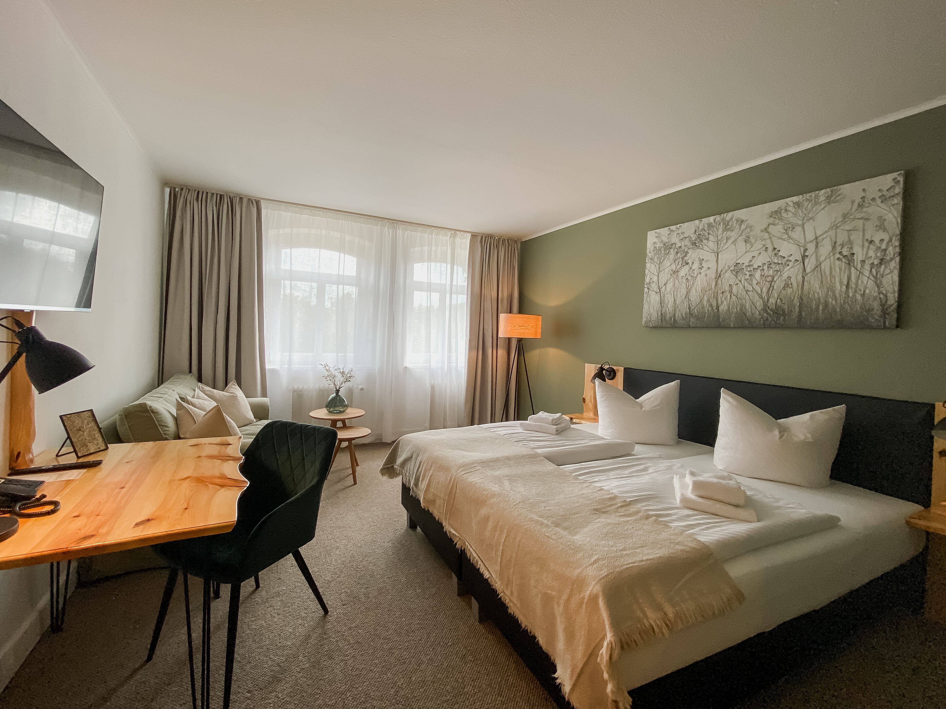 Une chambre d’hôtel avec un lit, un bureau, une chaise, une lampe, un vase de fleurs et un tableau au mur.