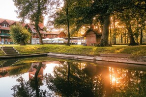 A serene outdoor setting with a reflective pond, a wooden deck, and a building with a red roof. - Waldhotel Eiche (Lübbenau/Spreewald)