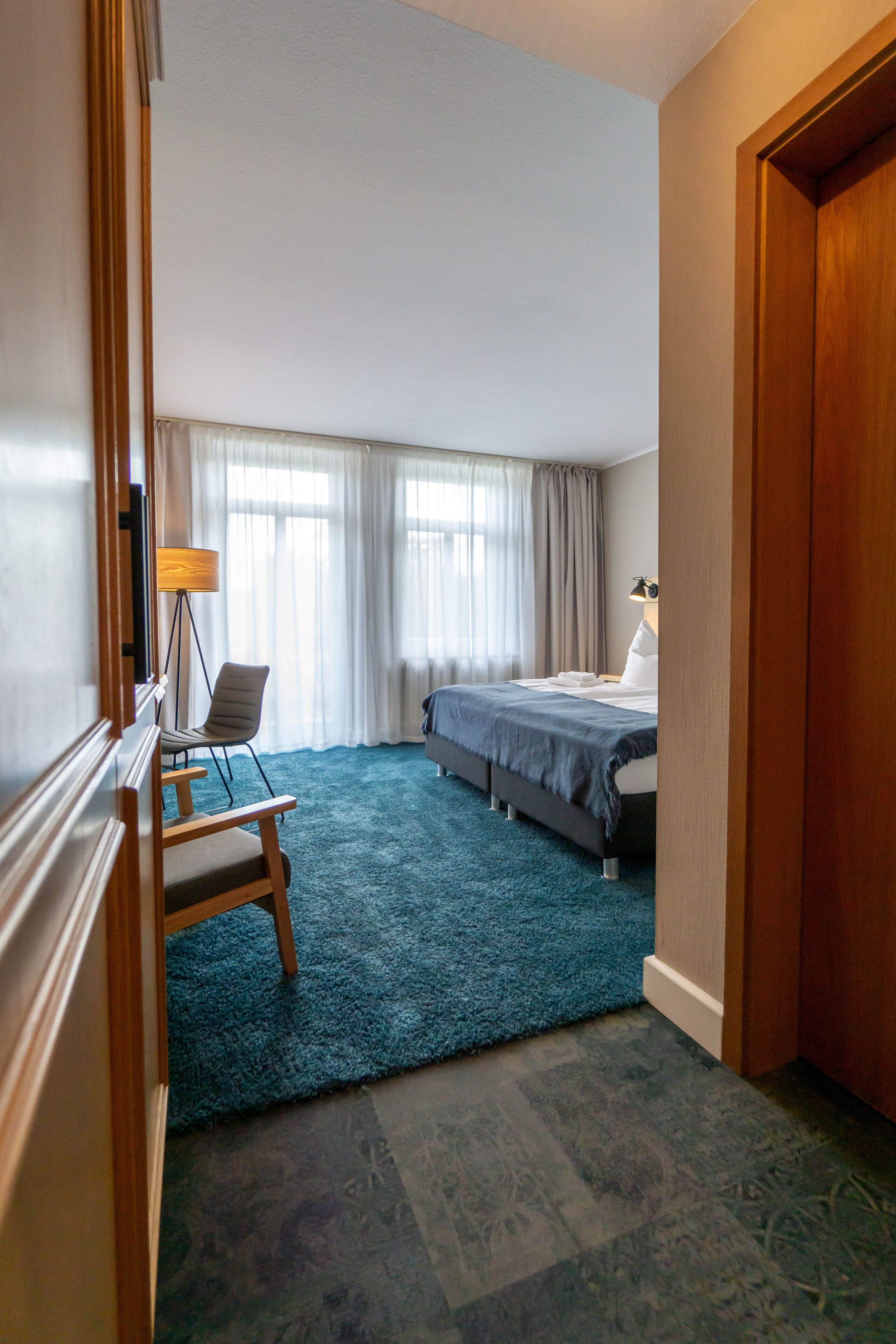 Habitación de hotel con cama, escritorio, silla, lámpara y una ventana grande con cortinas.