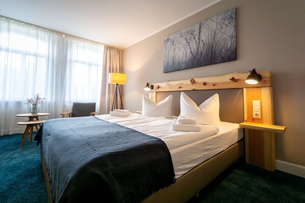 A hotel room with a bed, a bedside table with a lamp, a chair, a small table with a vase, and a painting on the wall. - Waldhotel Eiche (Lübbenau/Spreewald)