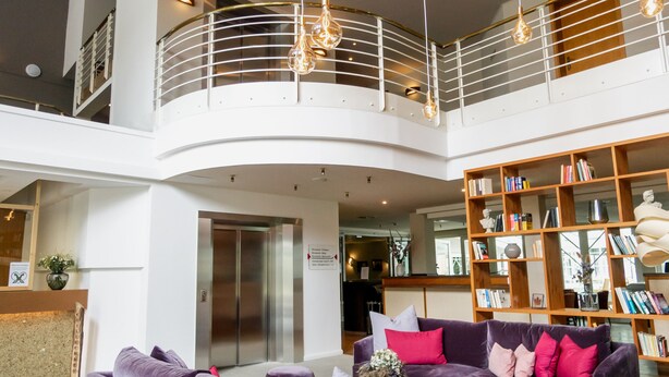 A spacious lobby with a high ceiling, a central seating area with purple sofas, a wooden bookshelf, and a modern chandelier.