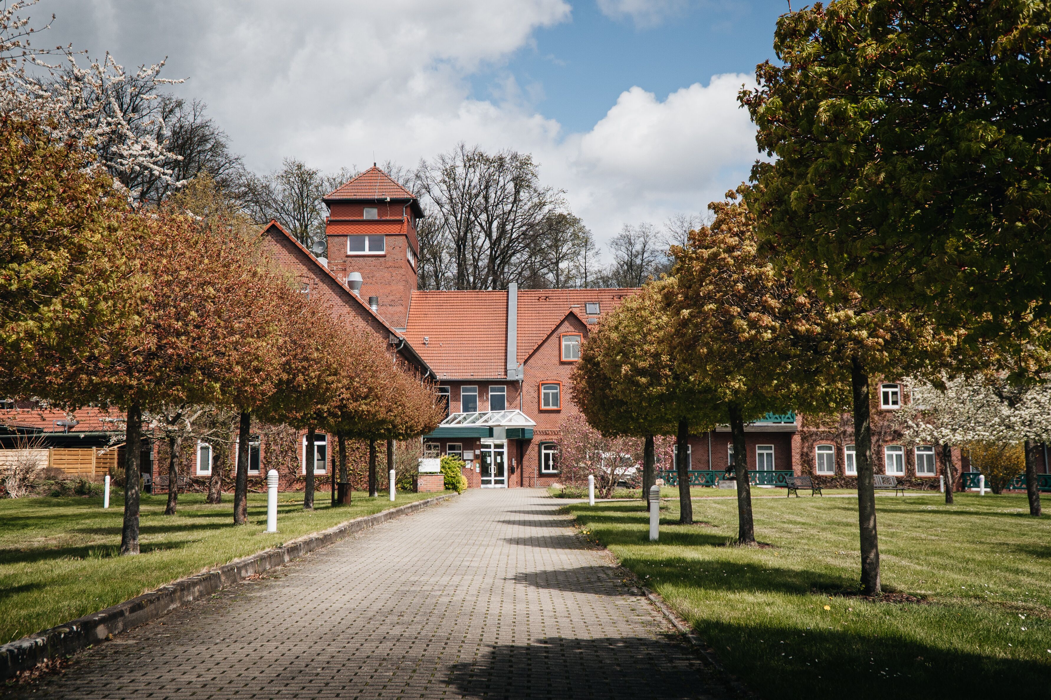 Un bâtiment en briques doté d’une tour centrale, entouré d’arbres et bordé d’une allée pavée.