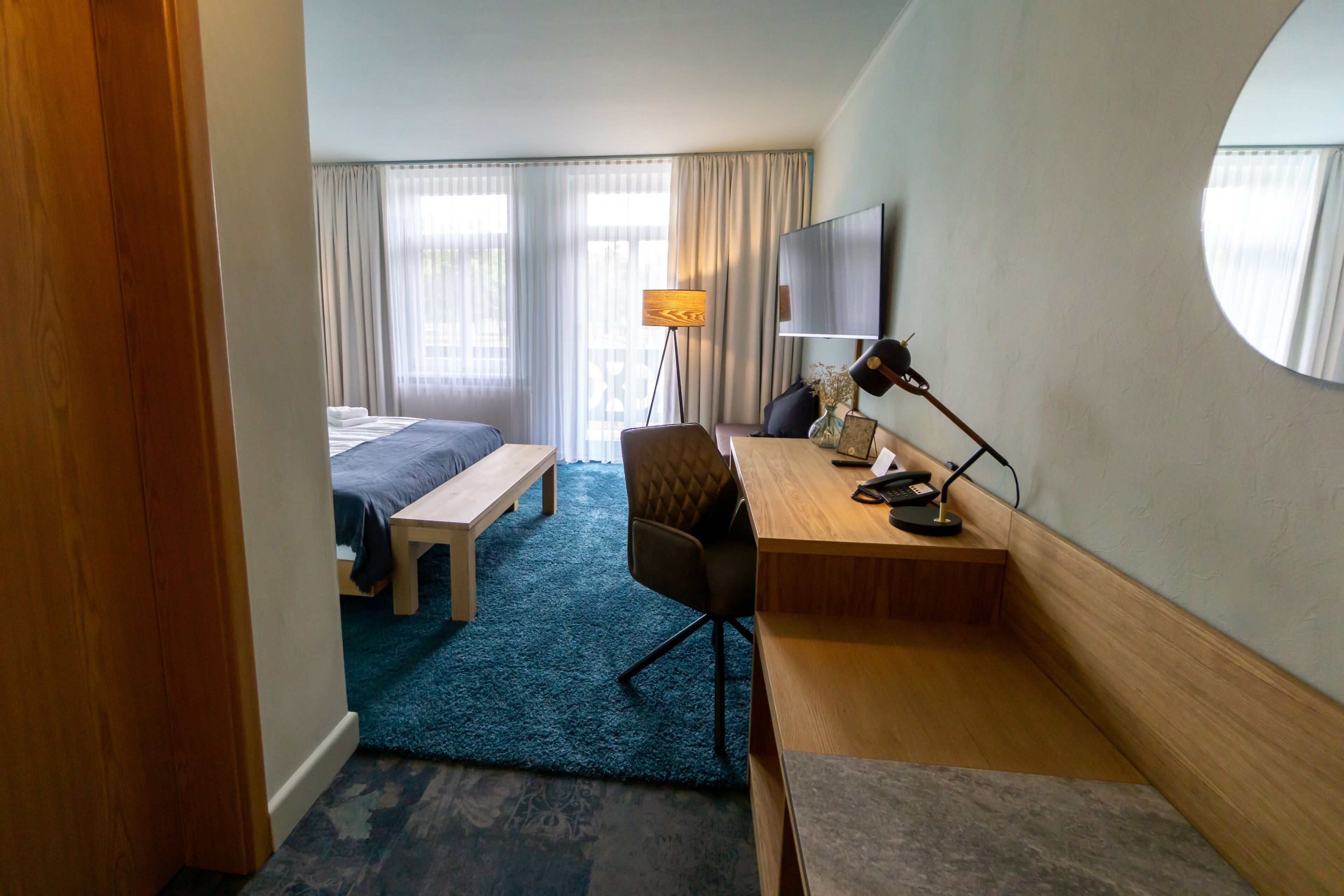 A hotel room with a bed, desk, chair, lamp, and a large mirror.