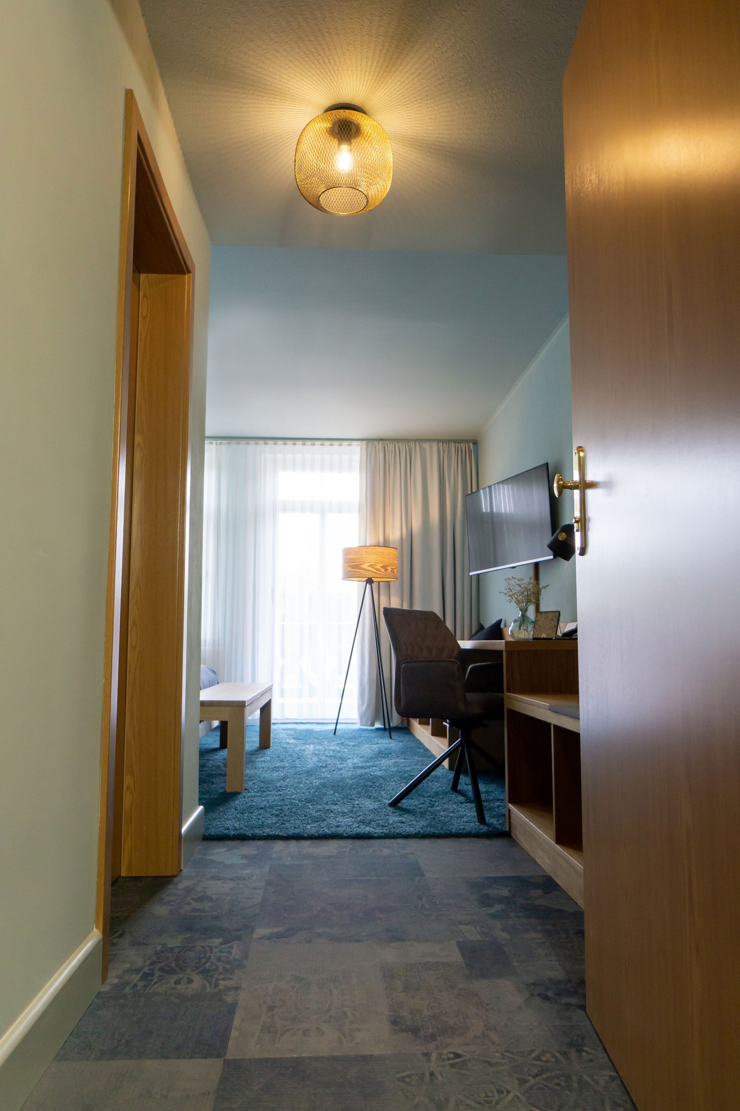 Et hotelværelse med skrivebord, stol, fjernsyn og et stort vindue med gardiner.