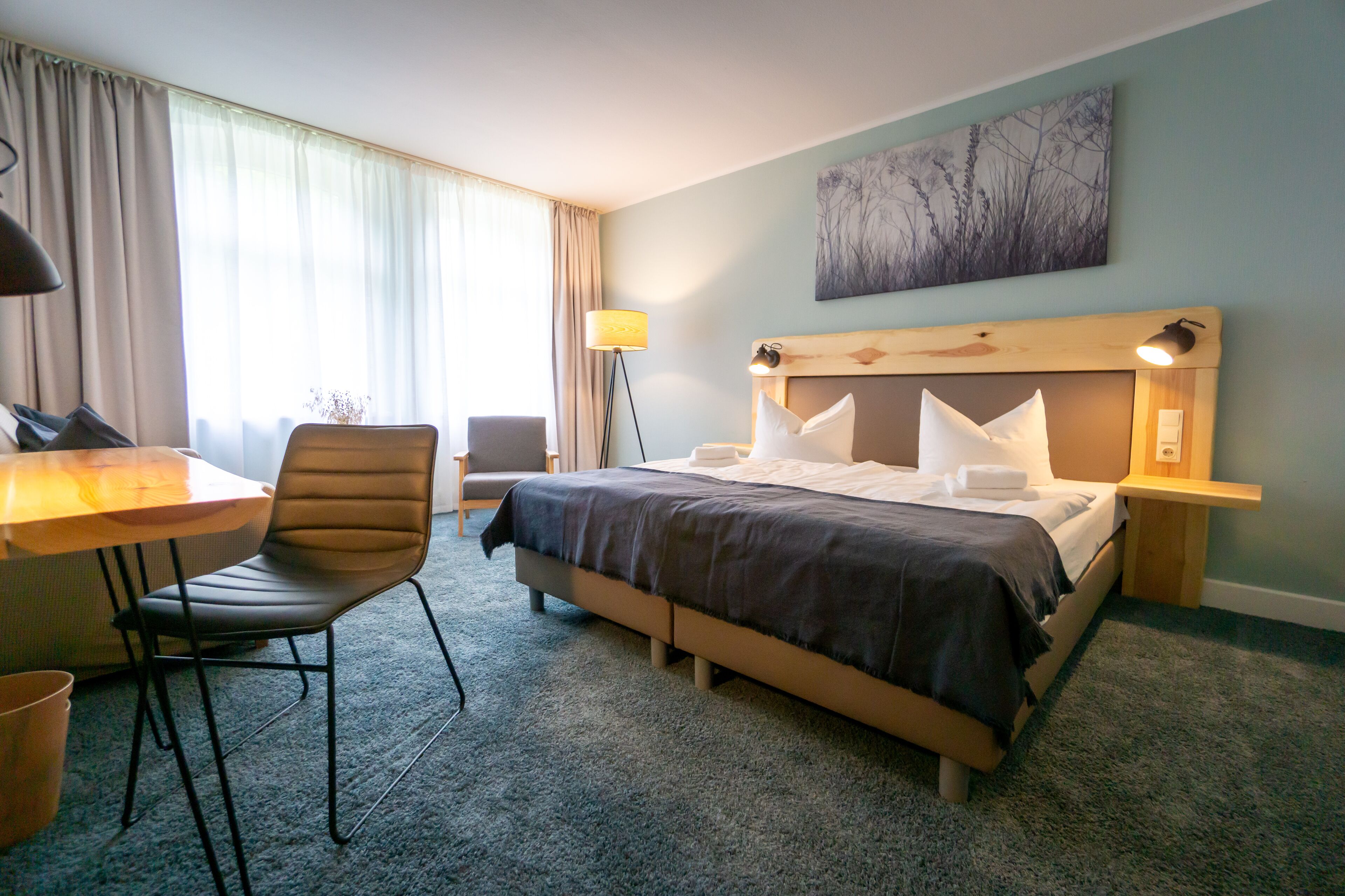 A hotel room with a bed, a desk, a chair, a lamp, and a painting on the wall.