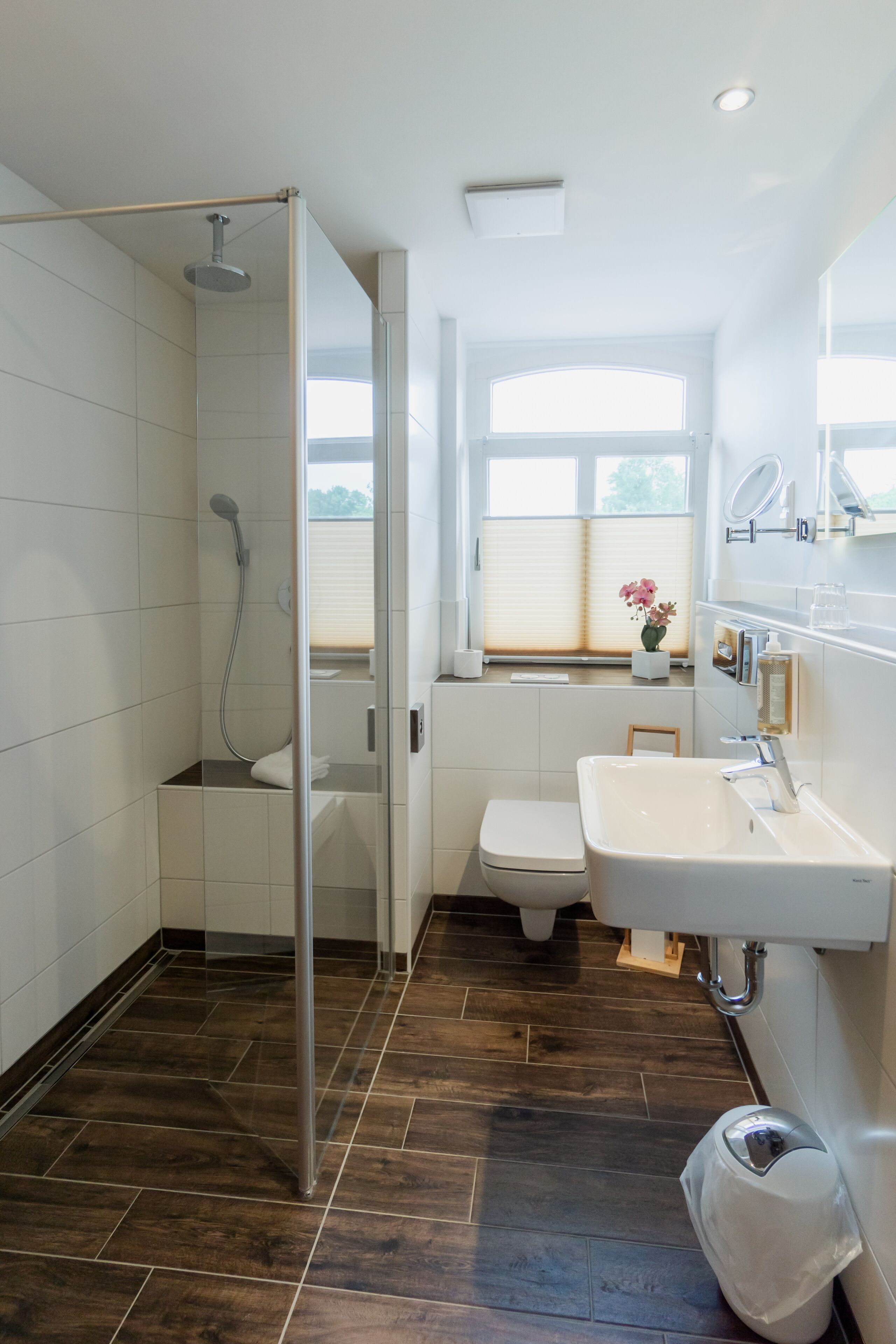 Ein modernes Badezimmer mit Duschkabine aus Glas, einer weißen Toilette, einem Waschbecken mit Spiegel und einem Fenster, das für natürliches Licht sorgt.