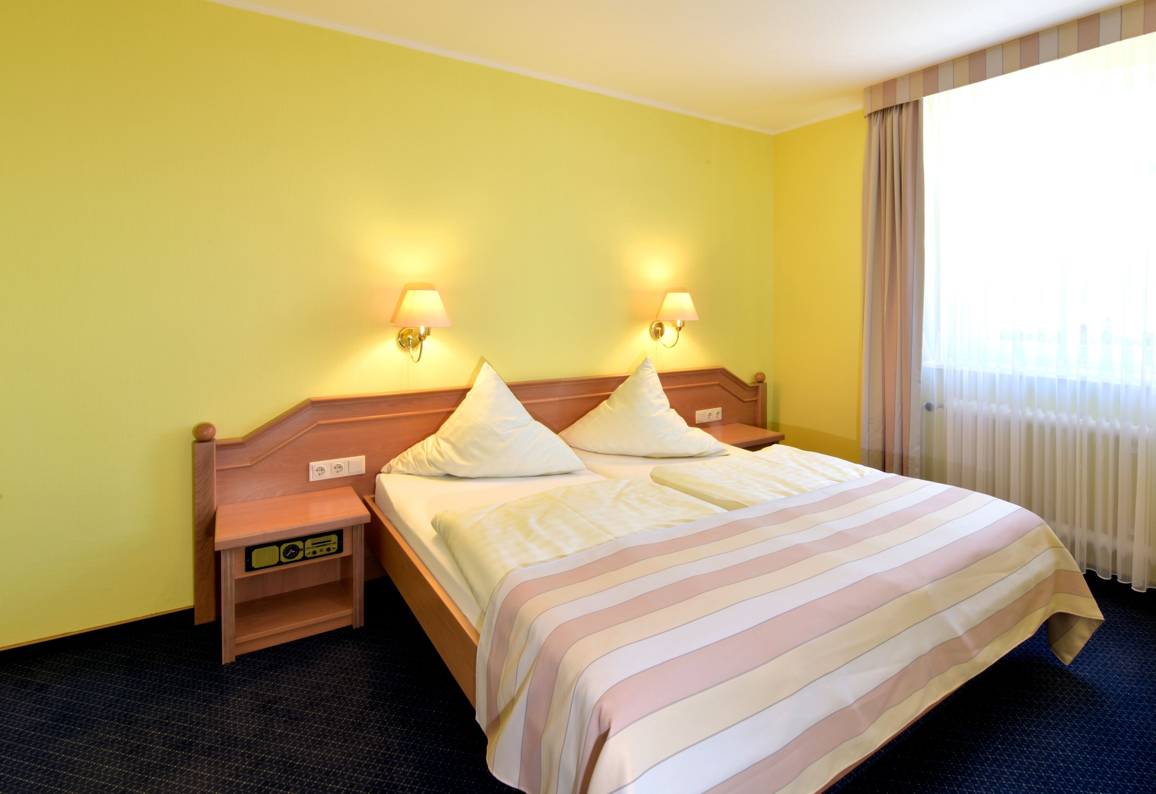 Una habitación de hotel con cama, dos lámparas de noche, una mesita de noche y una ventana con cortinas.