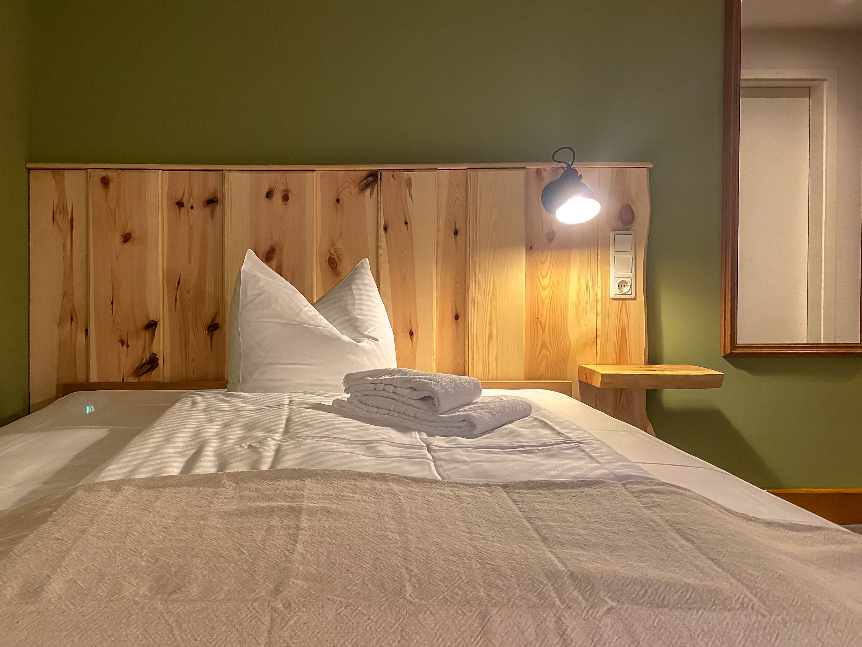Ein ordentlich bezogenes Bett mit weißen Leinen und Kissen, ein Holz-Kopfteil, ein Nachttisch mit einer Lampe und ein Spiegel an der Wand.