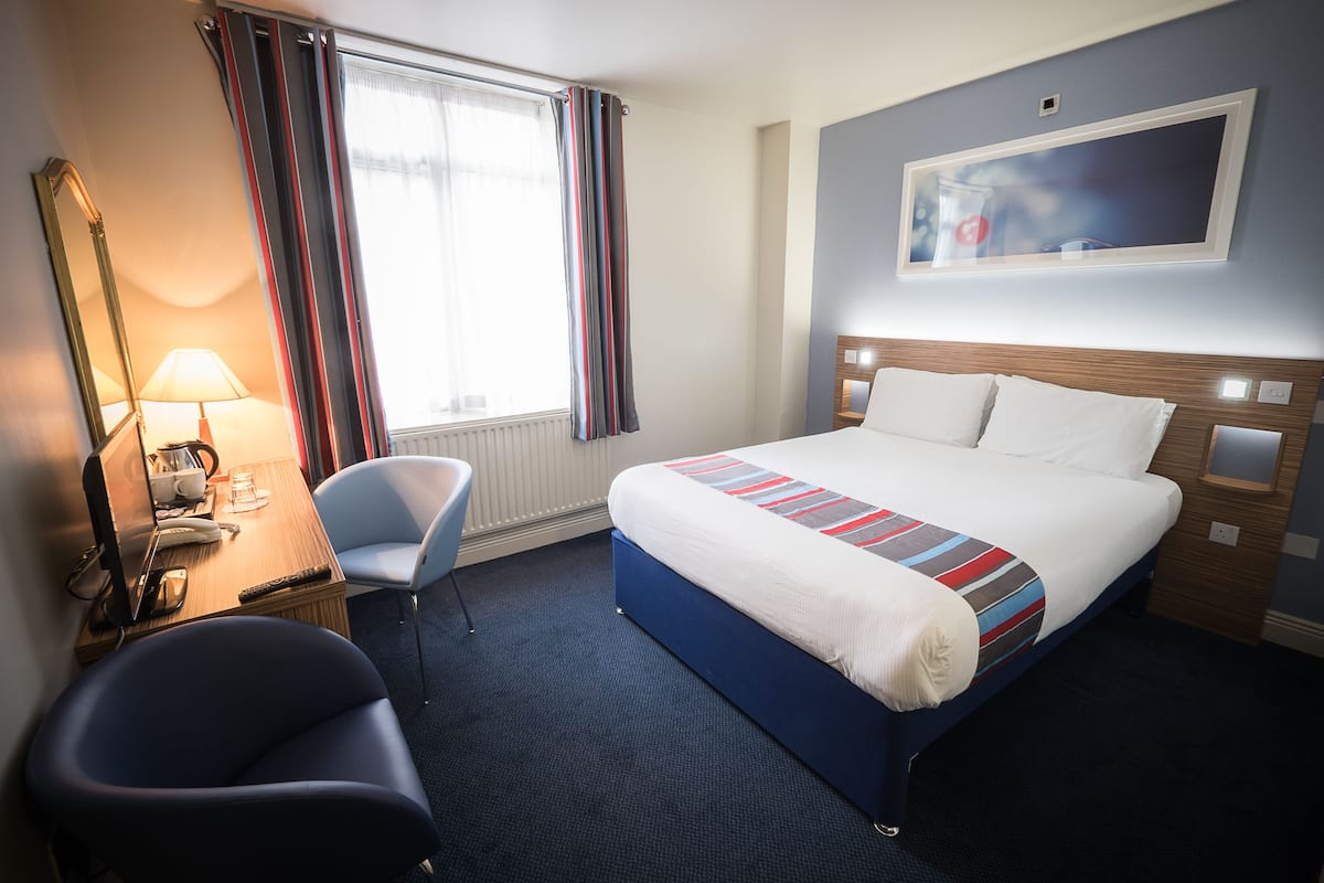 A hotel room with a bed, a desk with a computer, a chair, and a window with curtains.