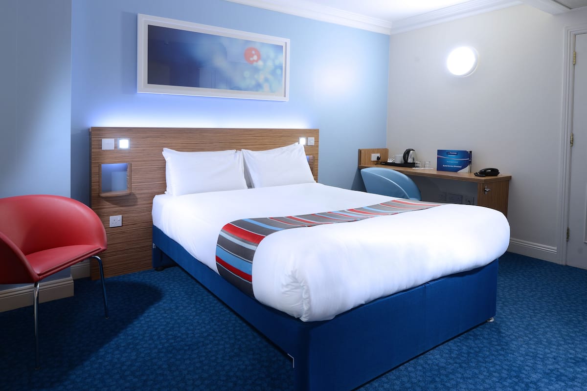 A hotel room with a neatly made bed, a red chair, a desk with a computer, and a painting on the wall.