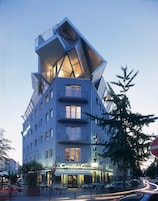 Un hotel moderno con un diseño angular distintivo, varios balcones y una entrada central con asientos al aire libre.