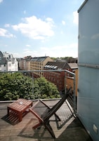 Una terraza en la azotea con una mesa y una silla de madera, con vistas a un paisaje urbano con edificios y árboles.