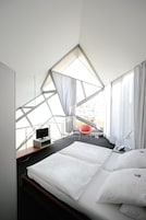 Una habitación de hotel moderna con una cama grande, un televisor sobre un mueble, una silla y vistas de la ciudad a través de amplias ventanas.