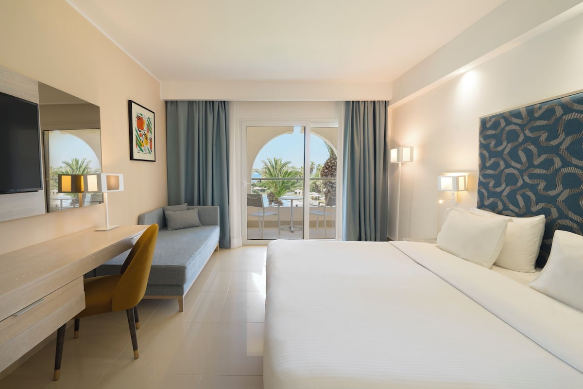 A hotel room with a bed, a desk, a chair, a TV, a balcony with a view, and a patterned headboard.