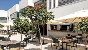 Un patio al aire libre con mesas, sillas, un bar y un gran árbol que da sombra.