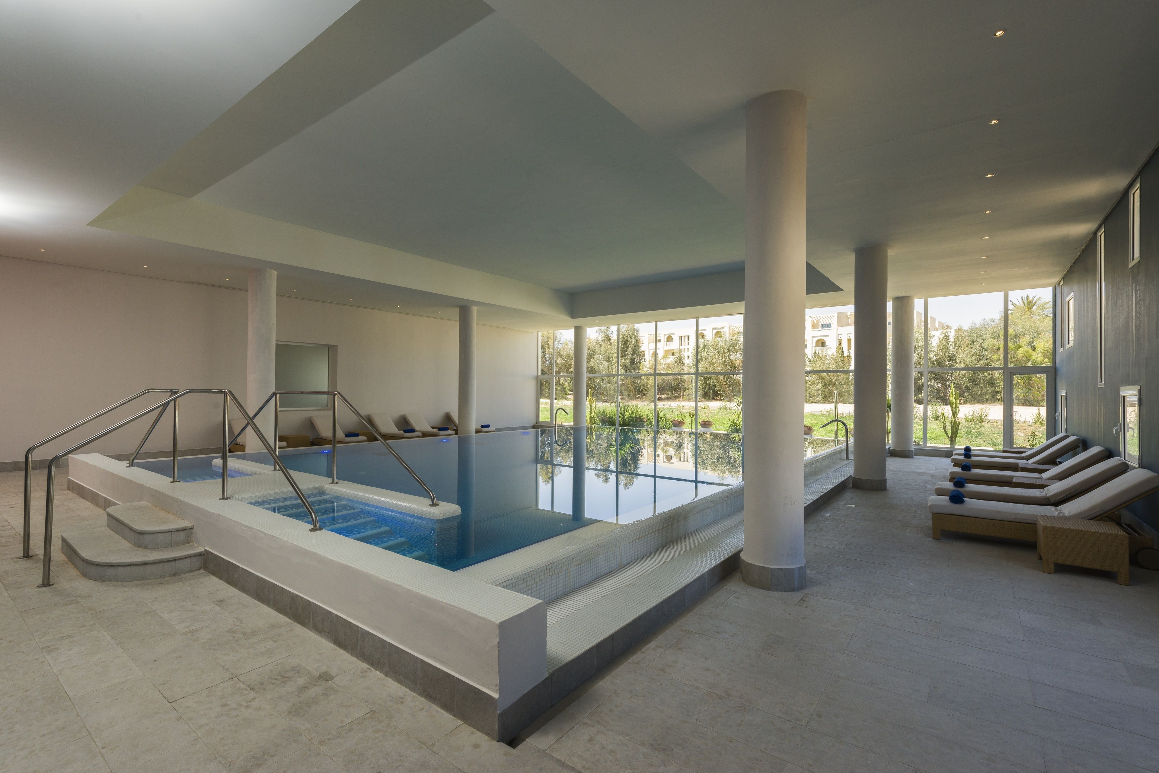 Uma ampla piscina interior com degraus para acesso, rodeada por espreguiçadeiras e grandes janelas que oferecem vista para o exterior.