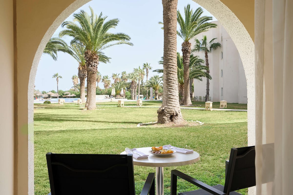 A view through an archway of a courtyard with palm trees, a table with a plate of food, and a building in the background.