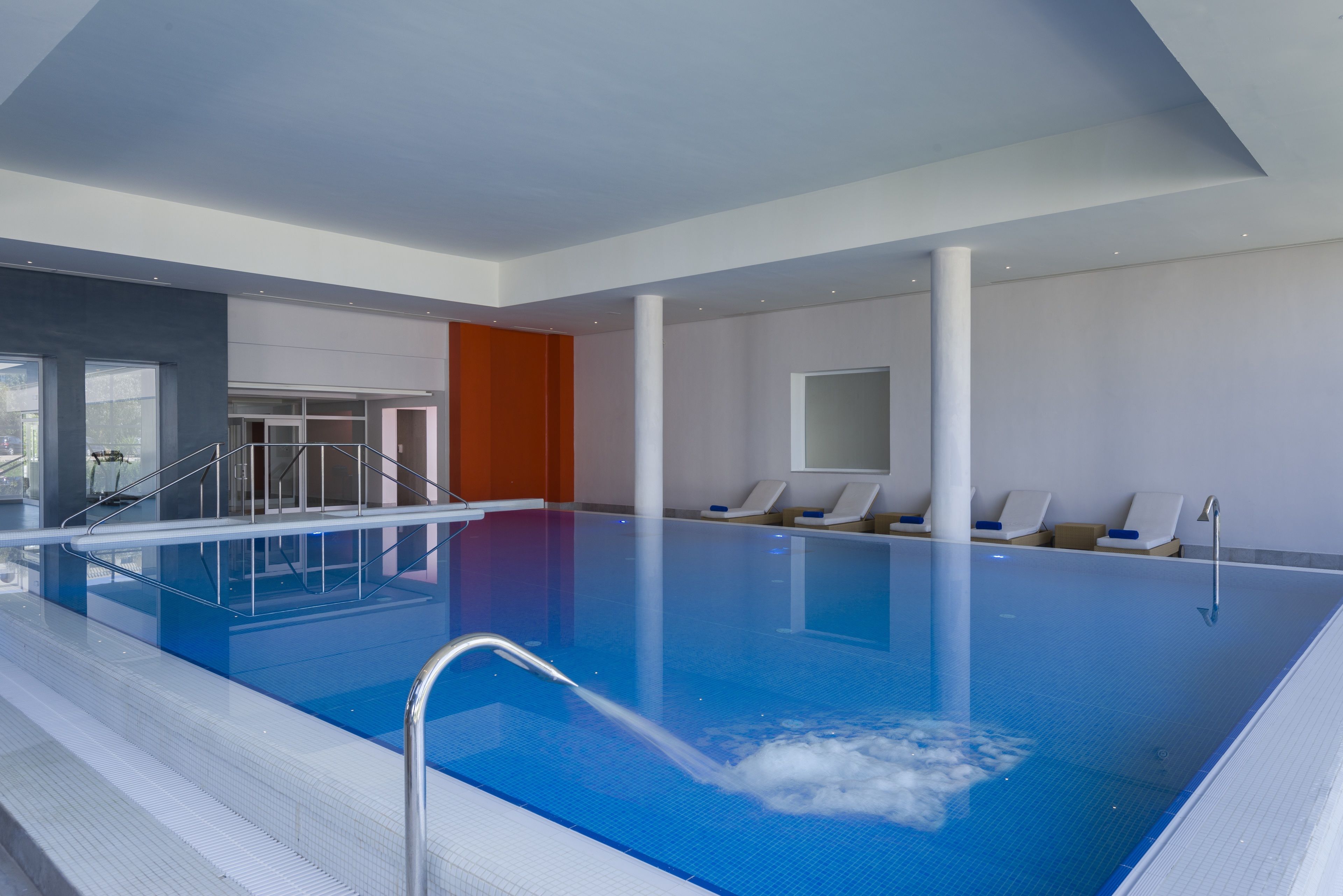 Una piscina interior con agua cristalina, un trampolín y sillones de descanso a un lado.